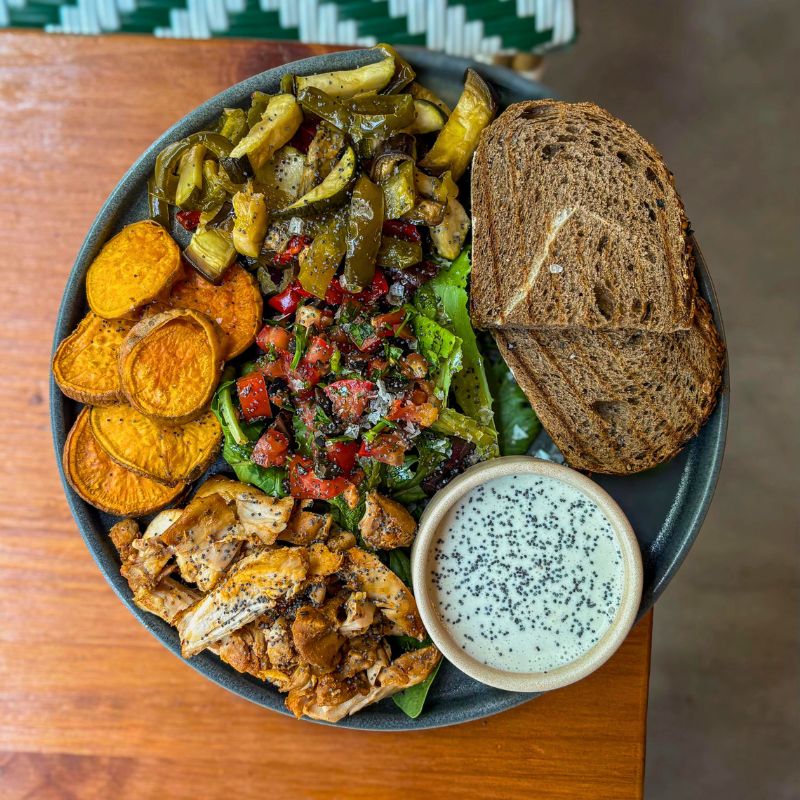 Plano contrapicado de plato de Mafrens Kitchen: pollo jugoso, tarrito de salsa de yogur, boniato asado, verduras al horno, ensalada en el centro y dos rebanadas de pan tostado de centeno.